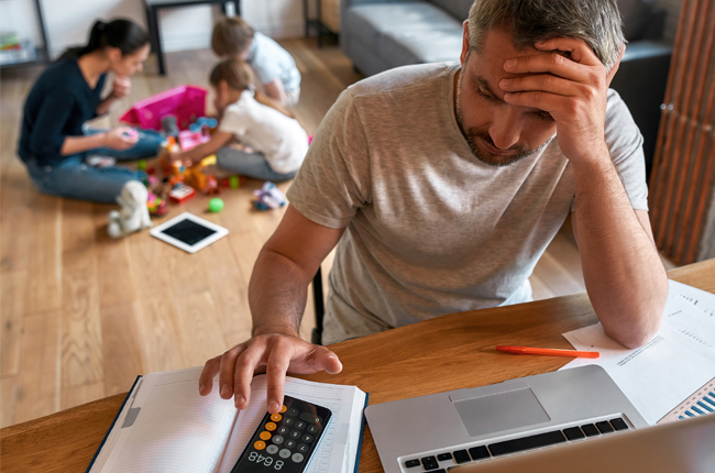 Man looking stressed while working on a laptop at home, with children playing in the background.